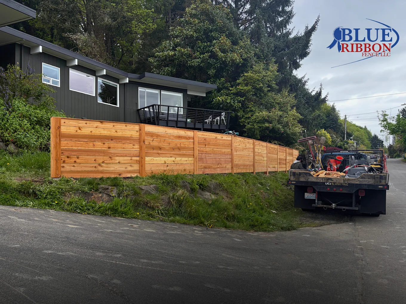 Long horizontal wood fence beside a dark gray home, with a Blue Ribbon work truck nearby.